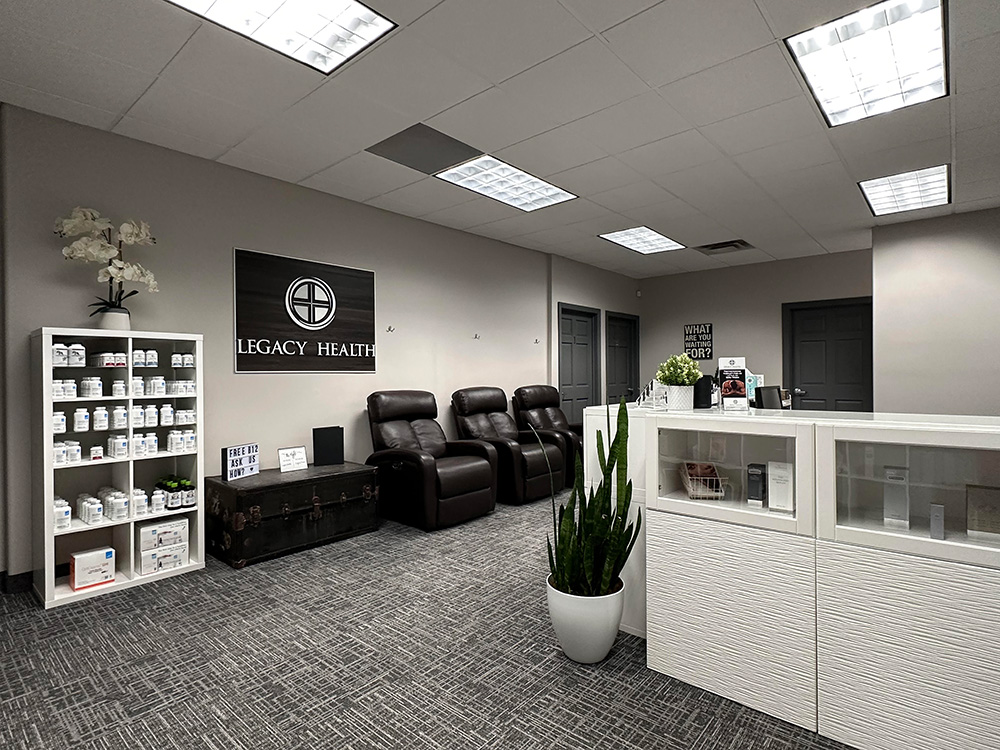 A modern medical office with a reception desk, a plant, and white storage cabinets on the right. On the left, there are shelves stocked with bottles and packages, a trunk, and three brown reclining chairs along the wall. A sign reads "About Legacy Health.