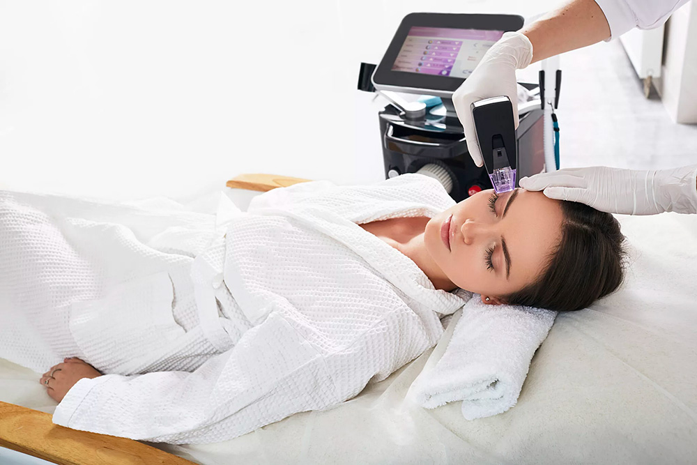 A person in a white robe lies on a treatment bed with their eyes closed while a professional wearing gloves uses the Morpheus8 handheld medical device on their forehead. A machine with a digital screen is visible in the background.