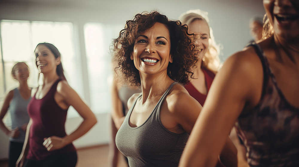A group of women are smiling and engaging in a lively exercise class in a well-lit room. Some are wearing workout tanks and tops, appearing to be in the middle of a fitness routine. The atmosphere looks energetic and joyful, providing an uplifting space for those navigating perimenopause and female hormones.