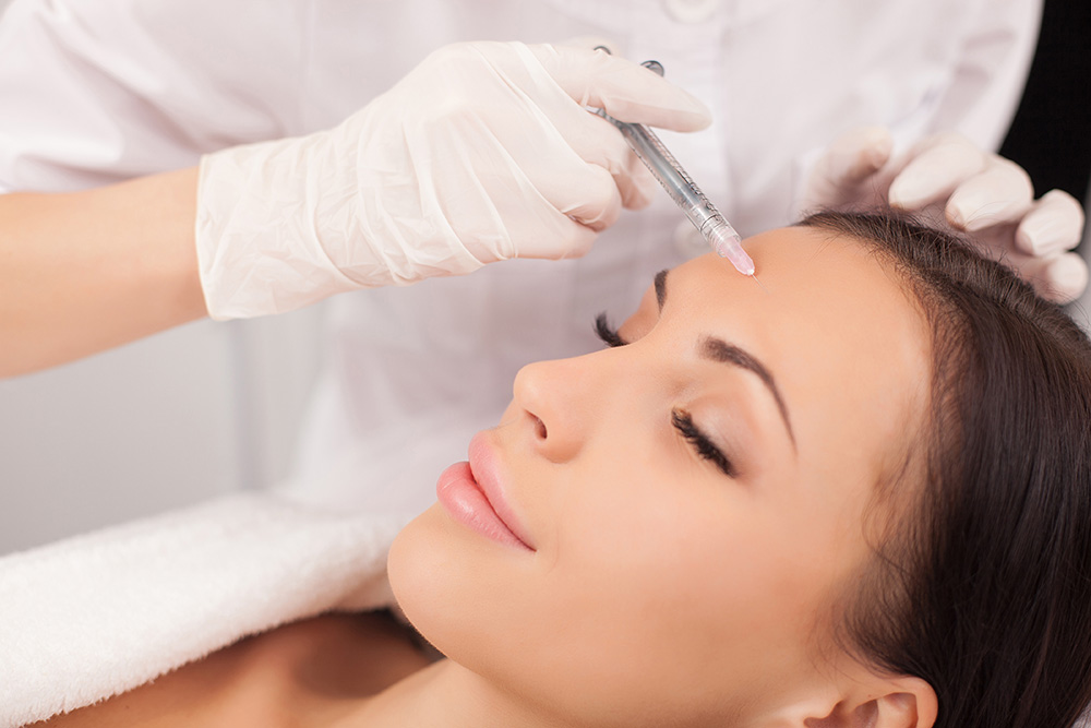 A person with closed eyes and a relaxed expression receives a beauty treatment injection in the forehead from a medical professional wearing white gloves. The setting, reminiscent of a serene med spa, exudes wellness and calm.