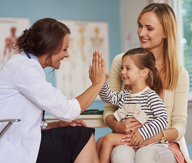 A doctor in a white coat gives a high-five to a smiling young girl sitting on her mother's lap. The mother, wearing a cream-colored cardigan, smiles at her daughter. The background shows anatomical charts on the wall, making the scene feel professional yet comforting.